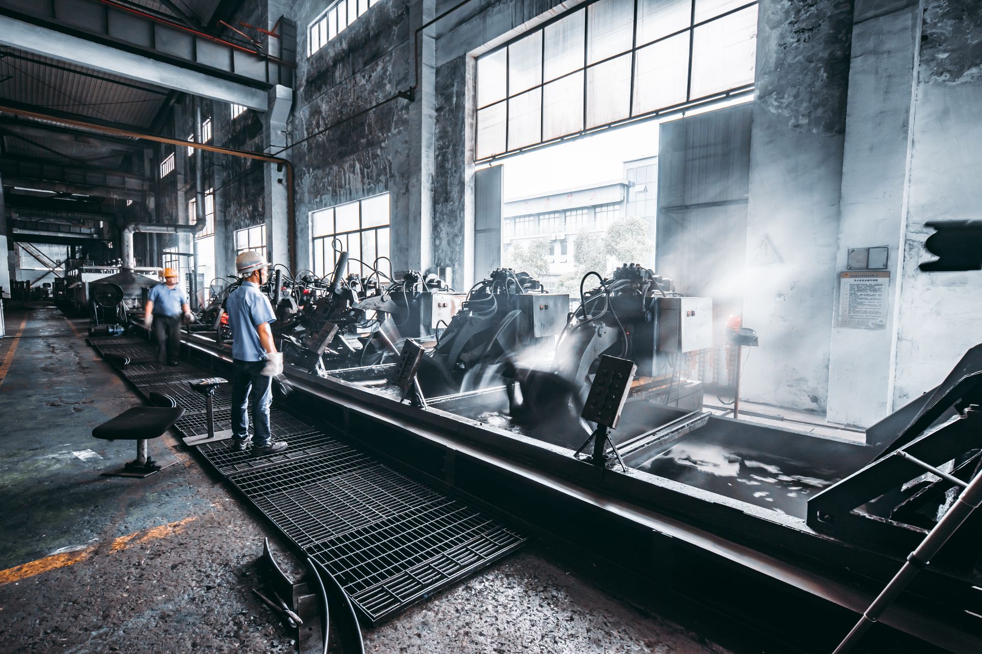 Factory interior with workers near machinery, steam, and large windows.