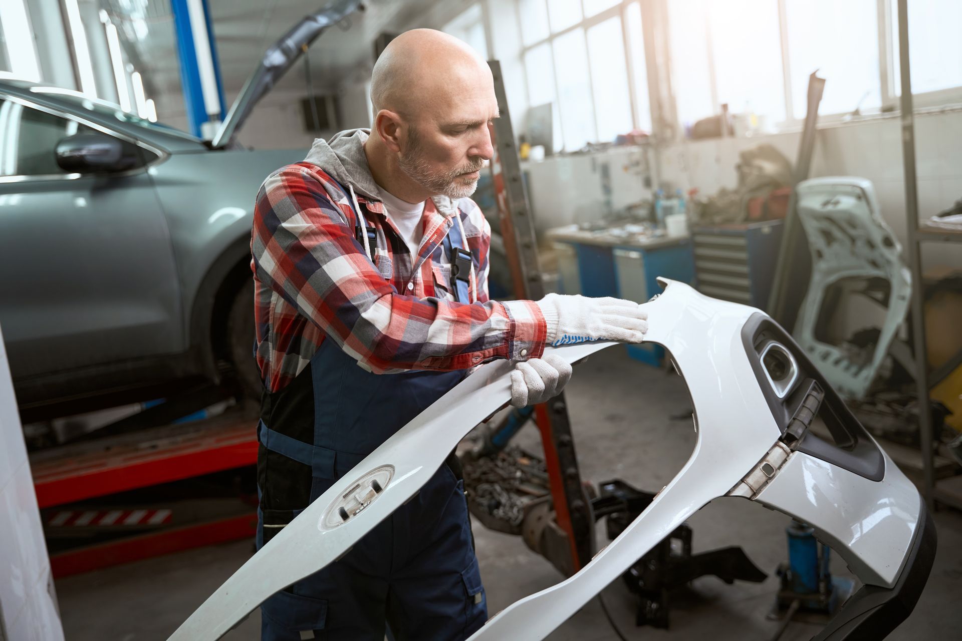 Mechanic in plaid shirt, wiping a white car bumper in a garage setting.