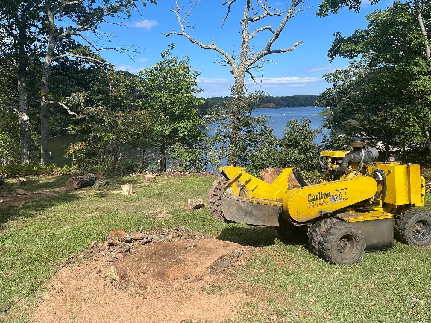 A yellow stump grinder is sitting in a grassy field next to a lake.