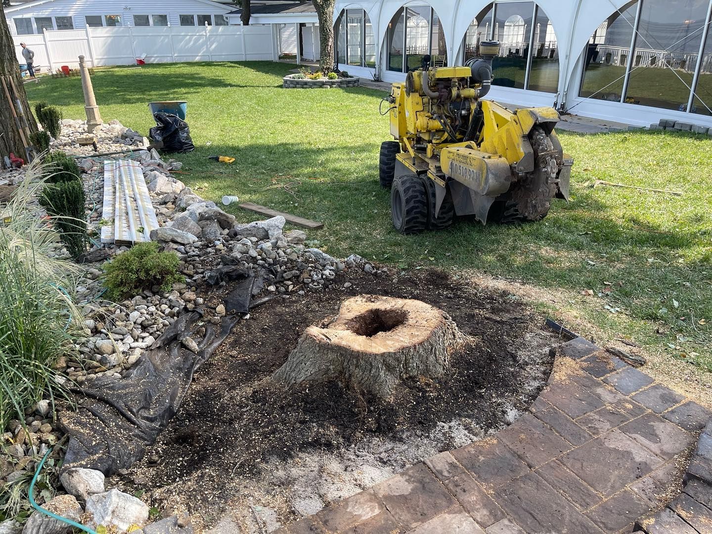 A yellow tractor is cutting a tree stump in a yard.