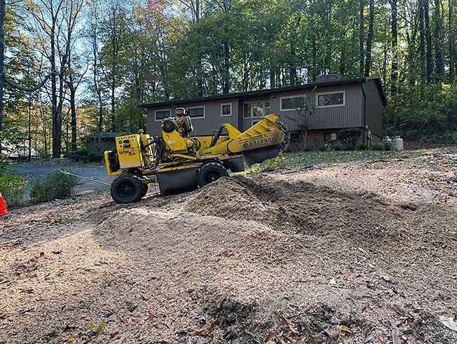 A yellow stump grinder is sitting in the dirt in front of a house.