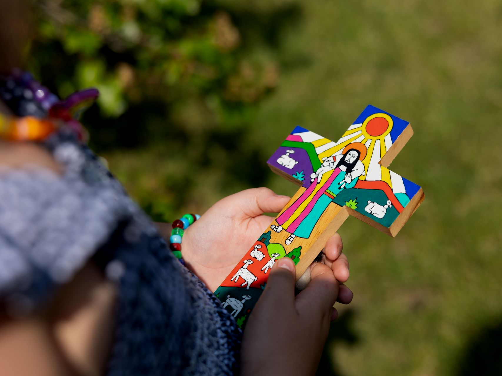 Hands holding a colorful painted wooden cross outdoors over a blurred green background
