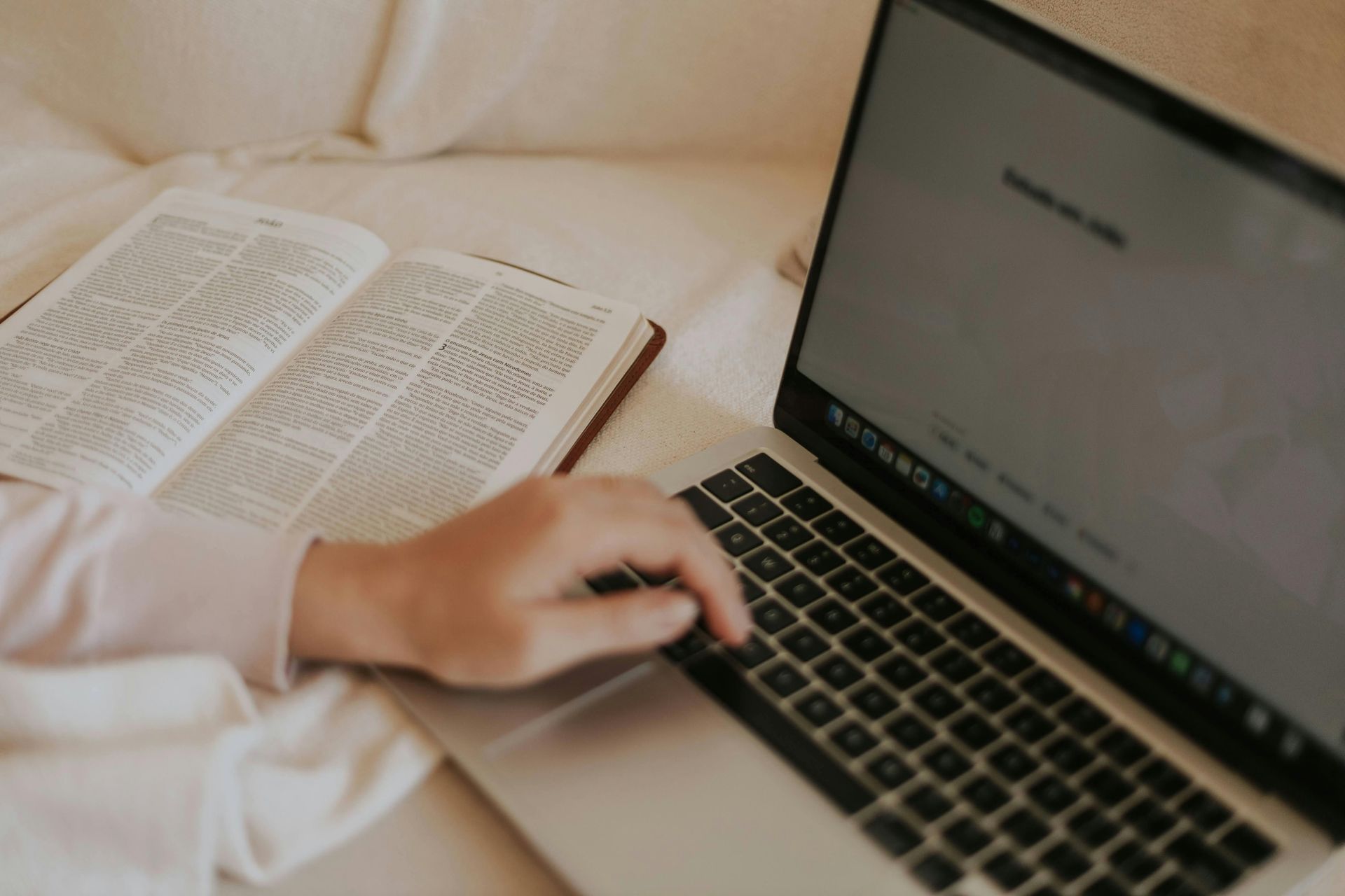 Hand resting on a laptop beside an open book on a bed