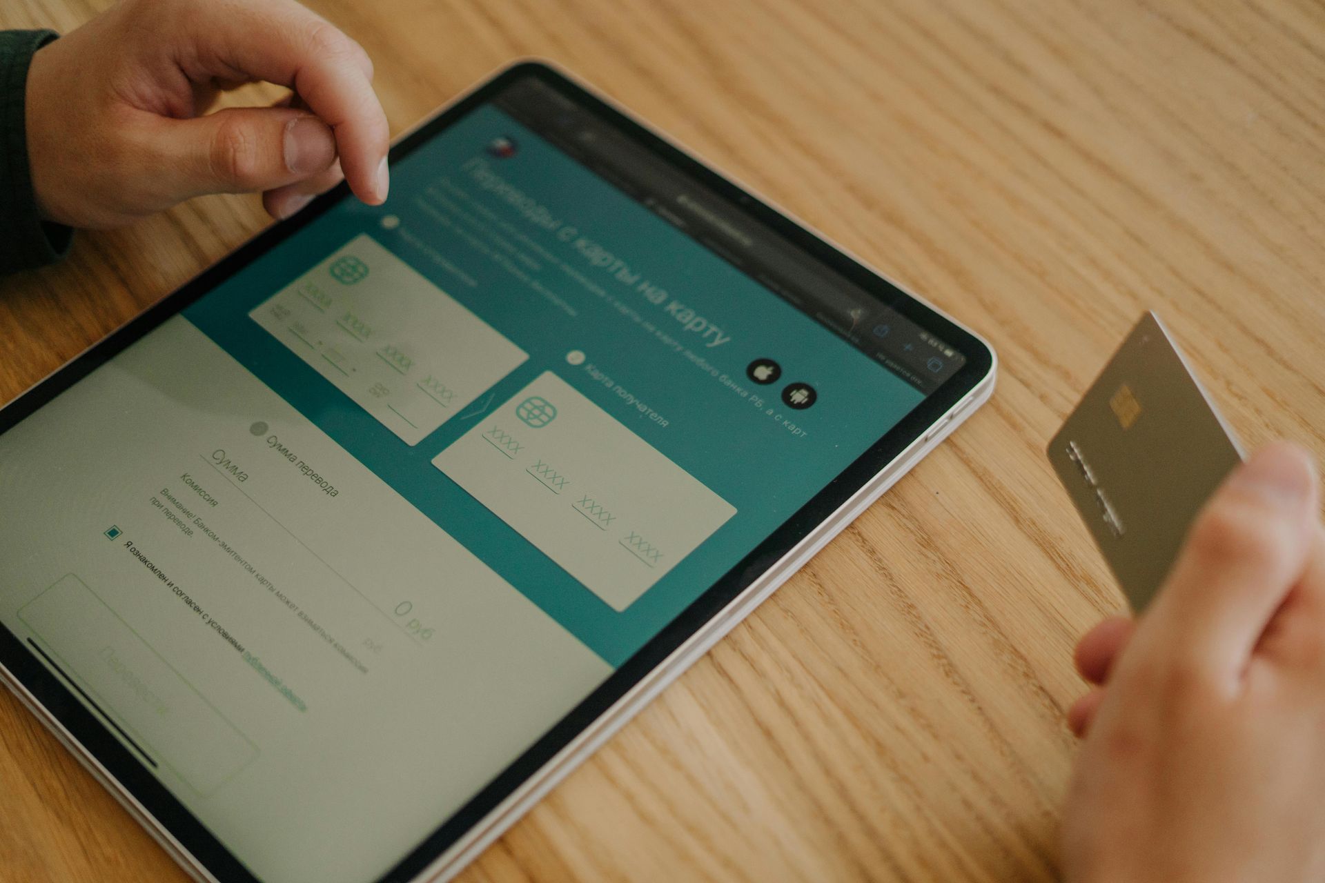 Hands holding a tablet and a card over a wooden table with a teal app screen.