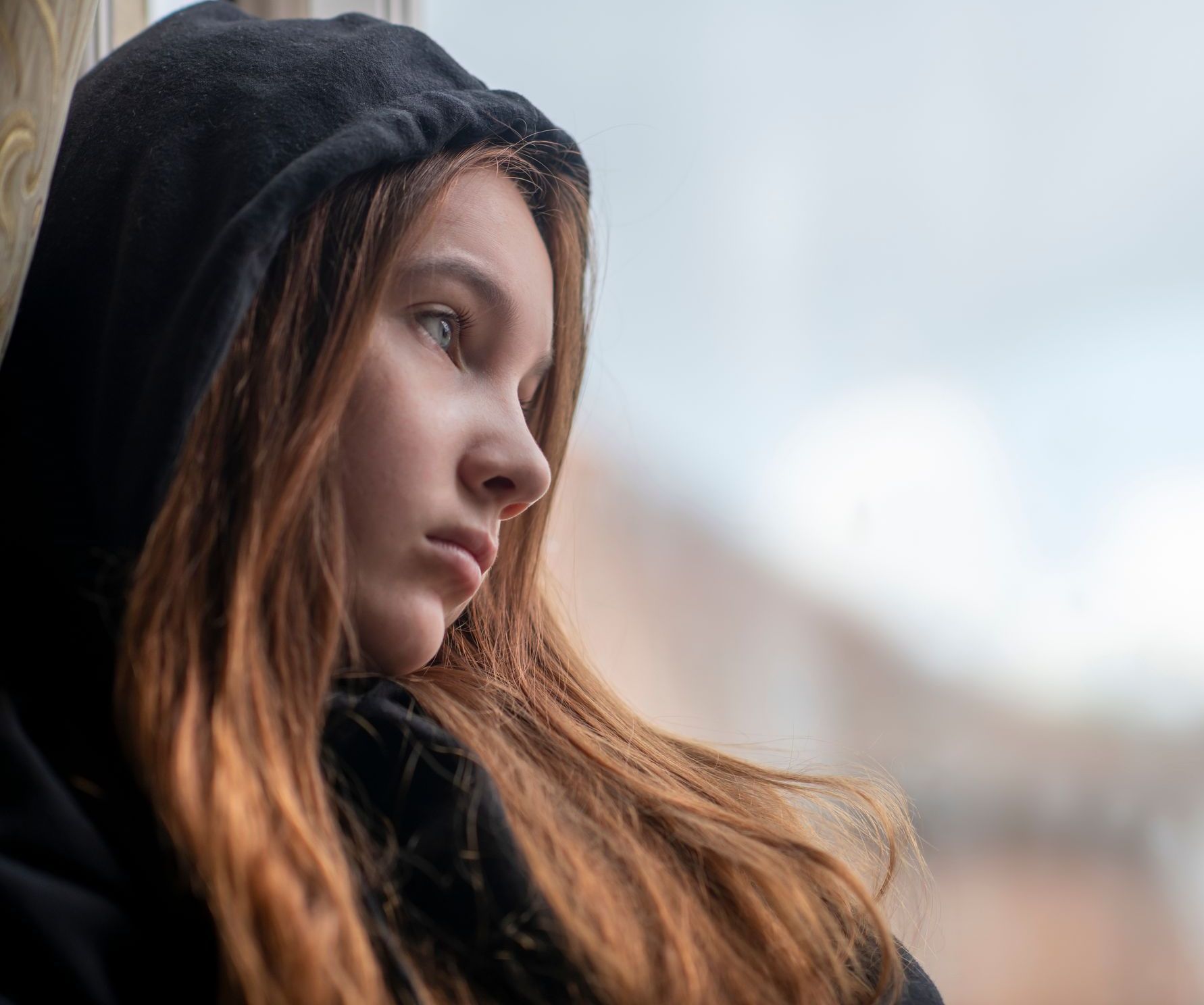 A young girl in a black hoodie is looking out of a window.