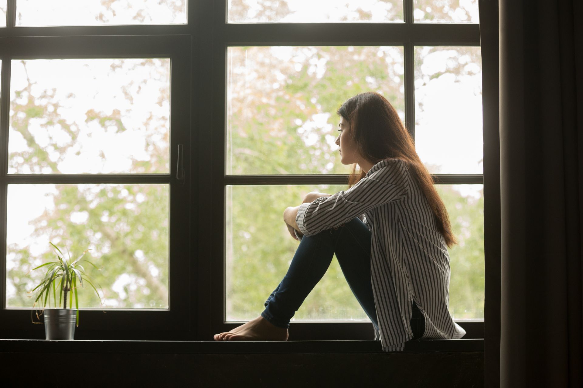 A woman is sitting on a window sill looking out the window.