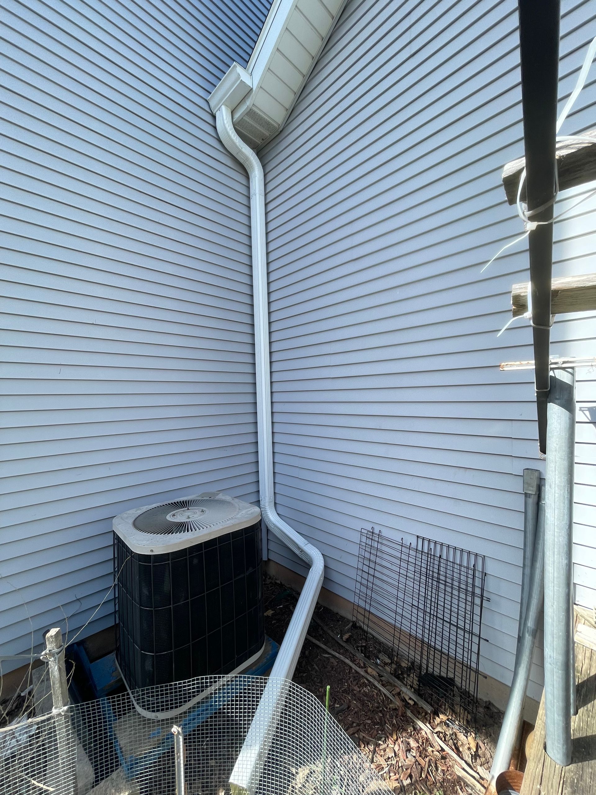A black air conditioner is sitting on the side of a white house next to a drain pipe.