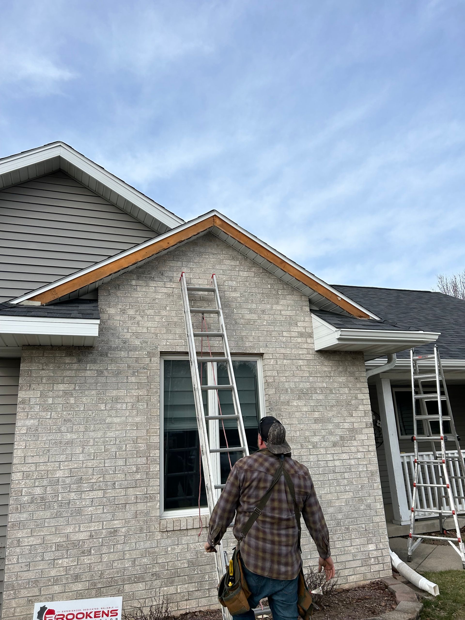 A man is standing in front of a brick house with a ladder.