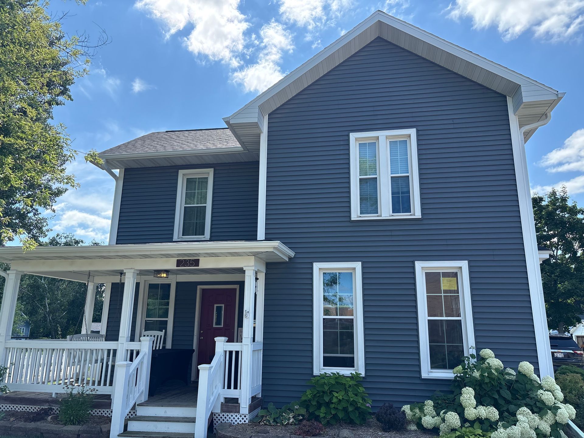 Two-story house with dark blue siding, white trim, and a porch, set against a partly cloudy sky.