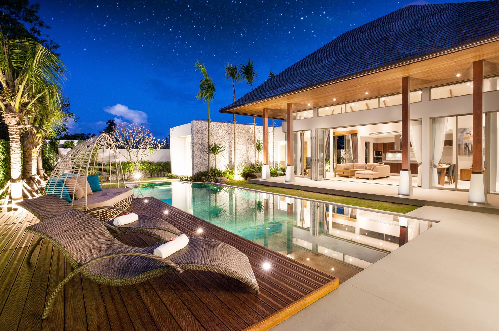 Luxury villa with a pool at night; lounge chairs, open doors to living area, starry sky.
