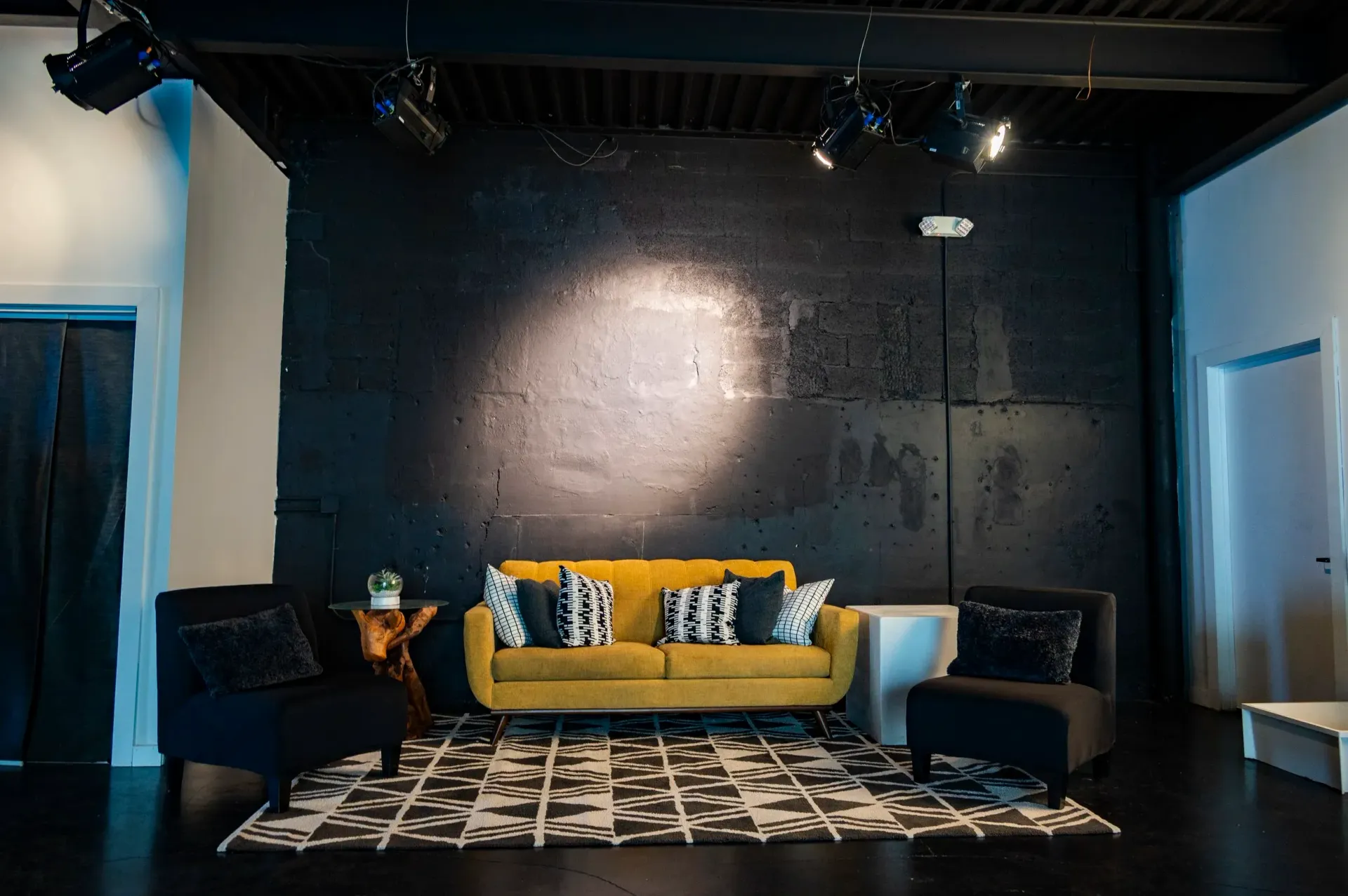 A yellow sofa and two black armchairs face a black wall in a modern studio. Spotlights shine down.