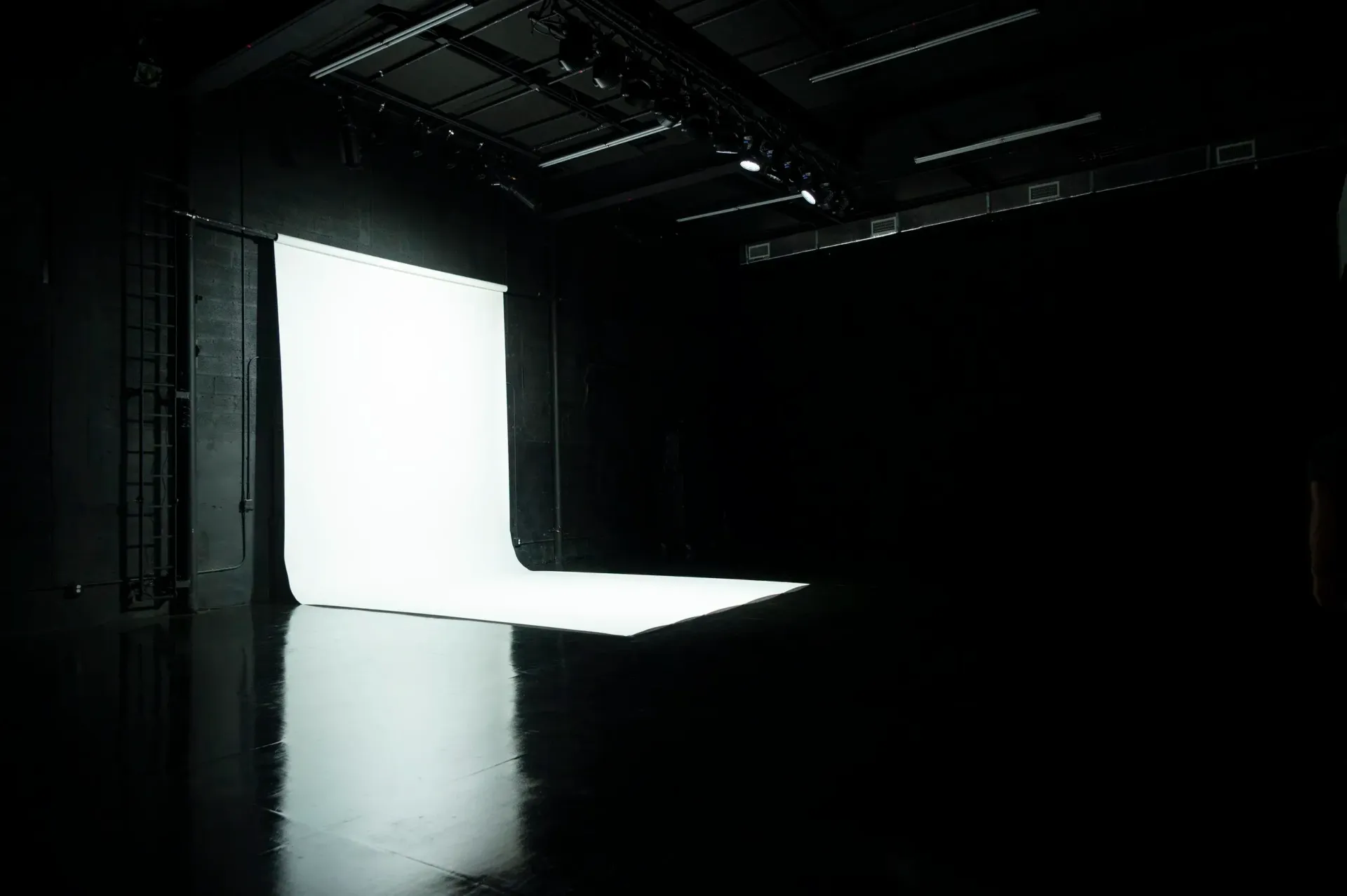 Empty, black studio with a bright white backdrop illuminated by overhead lights; shiny floor reflects the light.