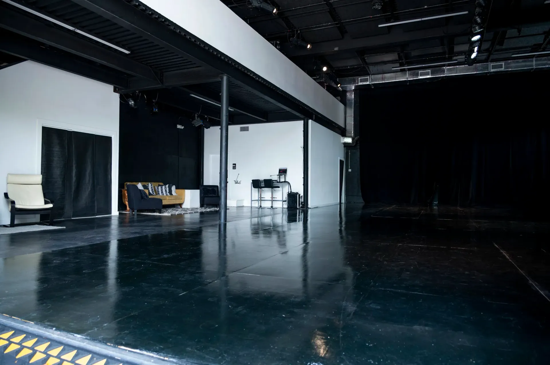 Empty theater stage with black floor and white walls. A piano and some furniture are visible.
