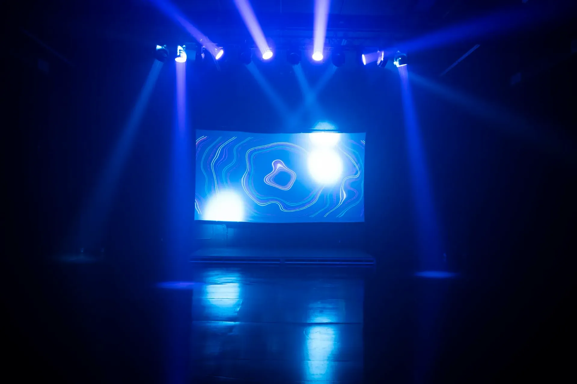 Stage bathed in blue light with a screen showing a swirling pattern; stage floor reflects the beams of light.