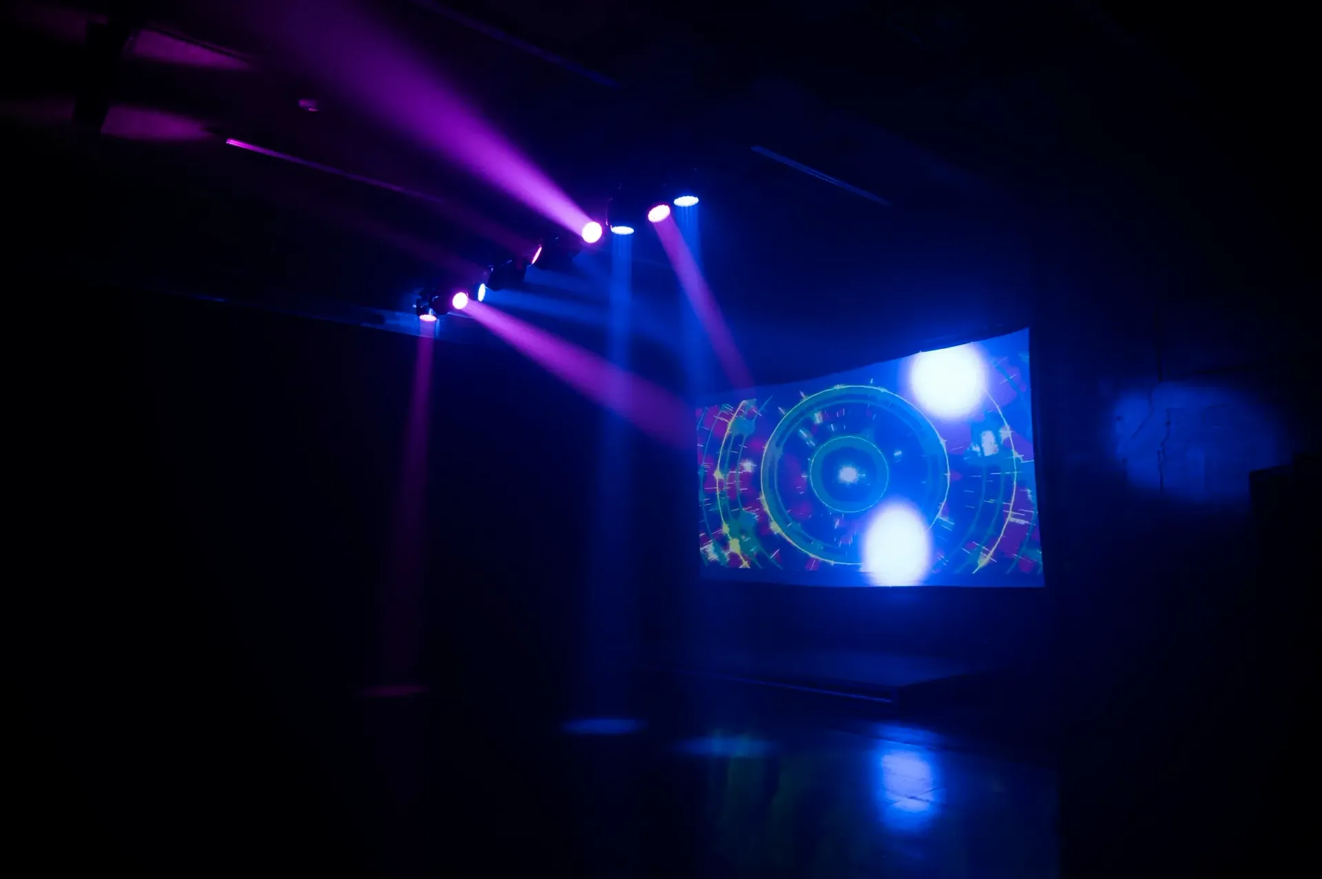 Dark room with purple and blue stage lights shining on a screen with a colorful, circular design.