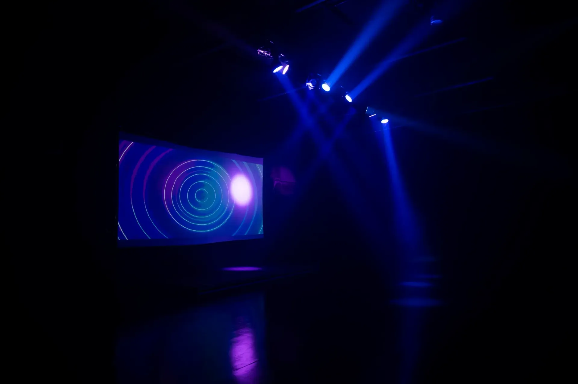 Dark room with a screen displaying a purple and white spiral pattern; lit by bright blue spotlights.