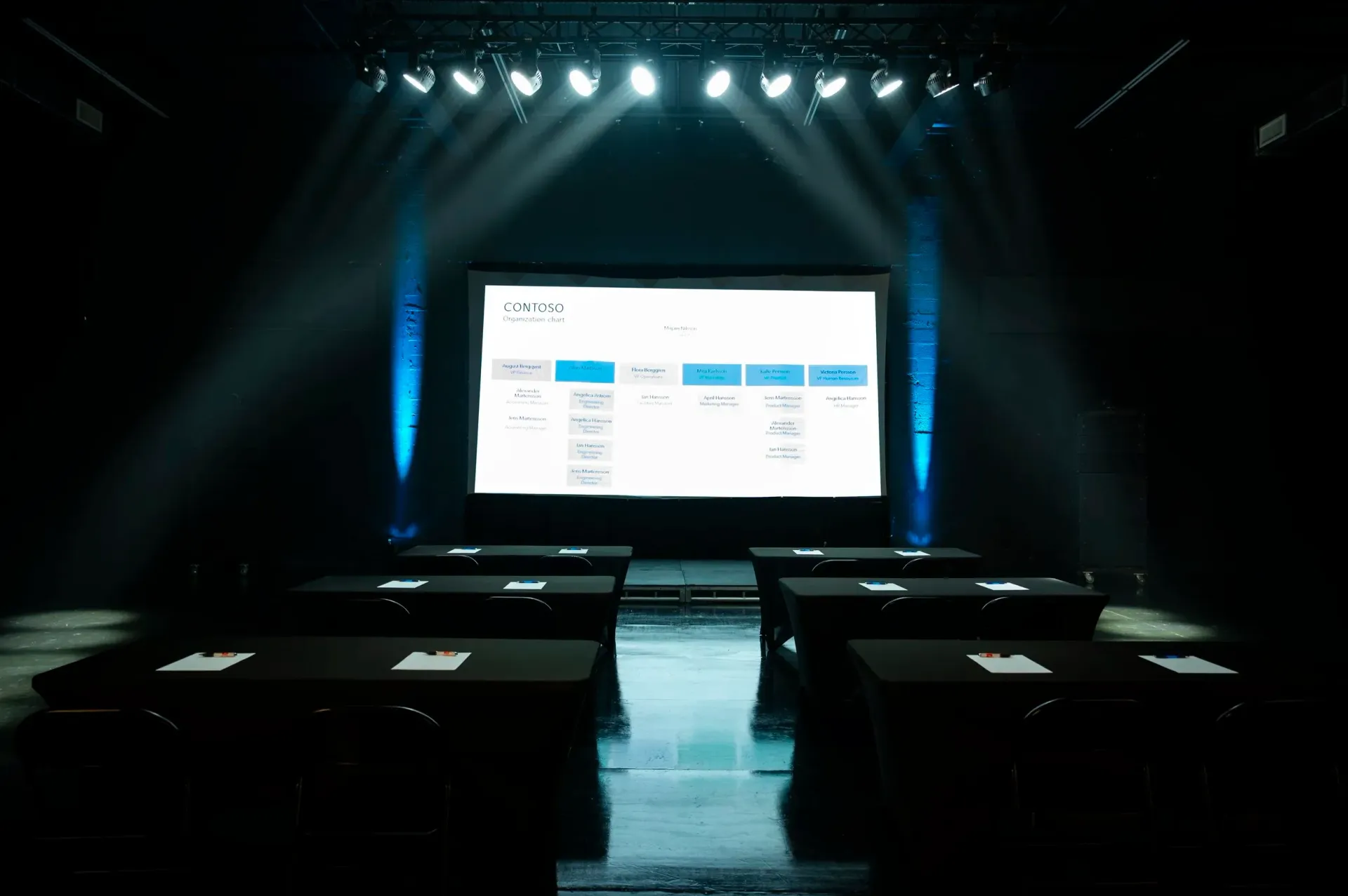 Empty stage with tables facing a large screen displaying information, under bright spotlights.