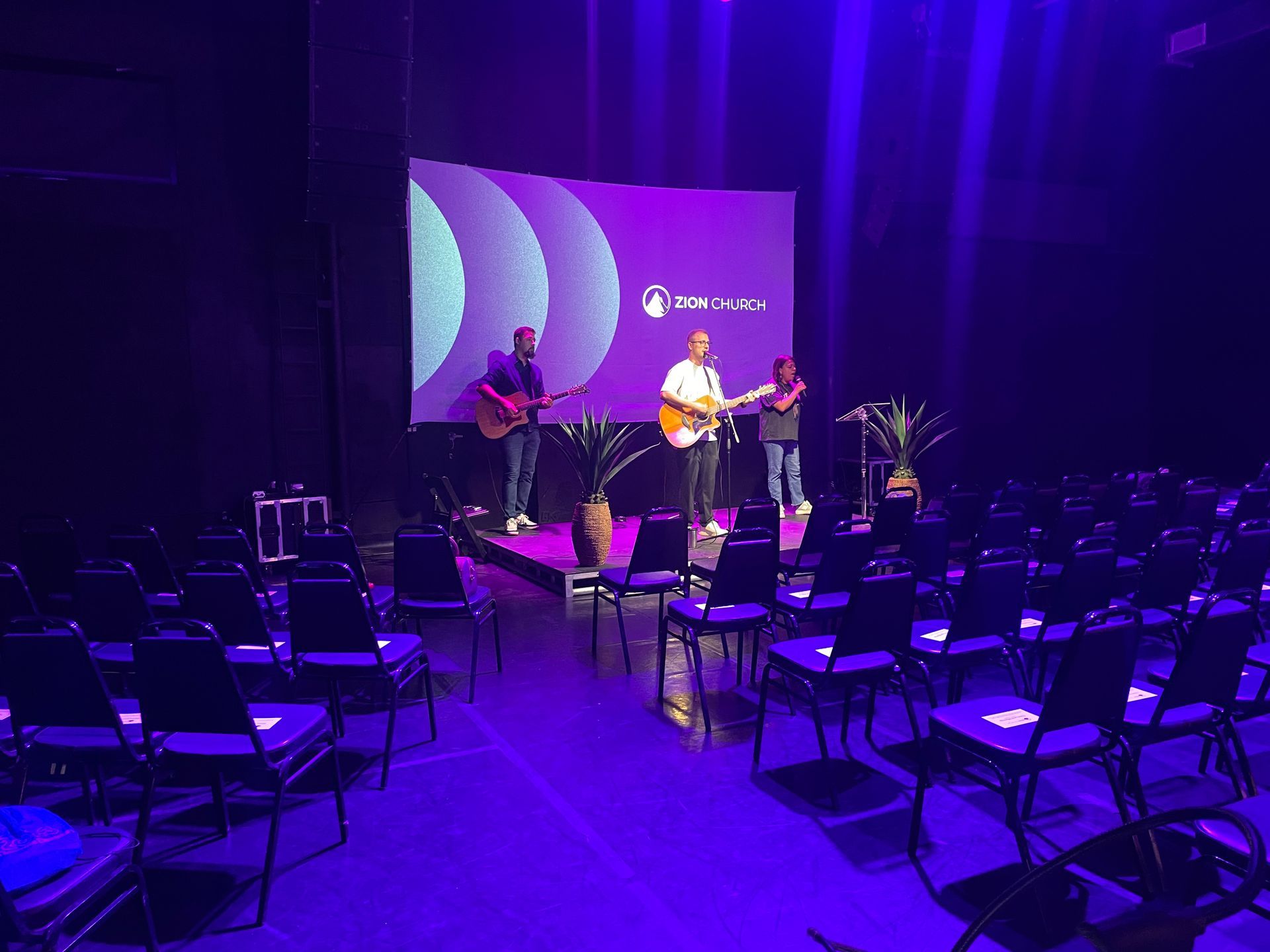Three musicians on stage playing guitars, with purple lighting and a screen behind them.