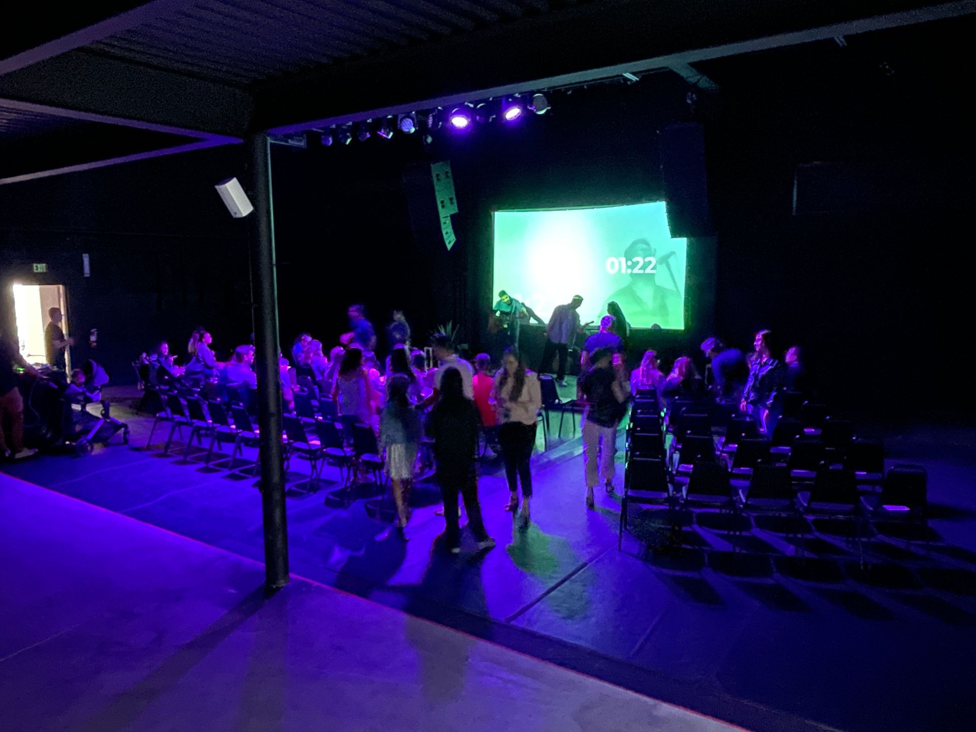 People in a dark venue with purple lighting. Rows of chairs face a lit stage with a projected image.