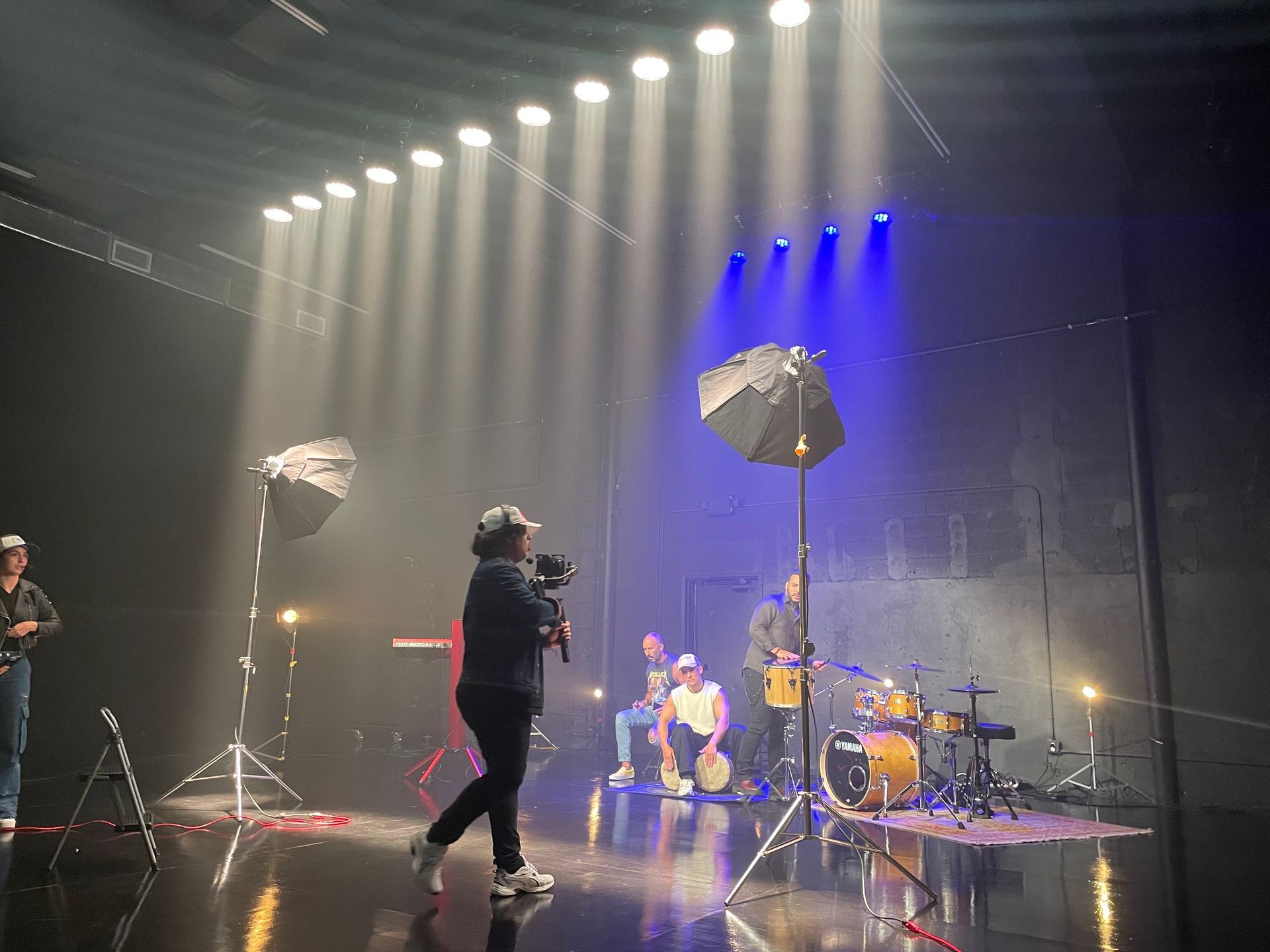 Band on stage under spotlights, filming a music video. Crew members and equipment visible.