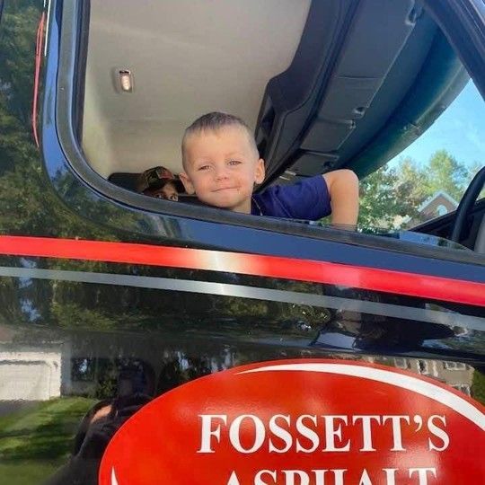 A boy is sitting in the driver 's seat of a fossett 's asphalt truck