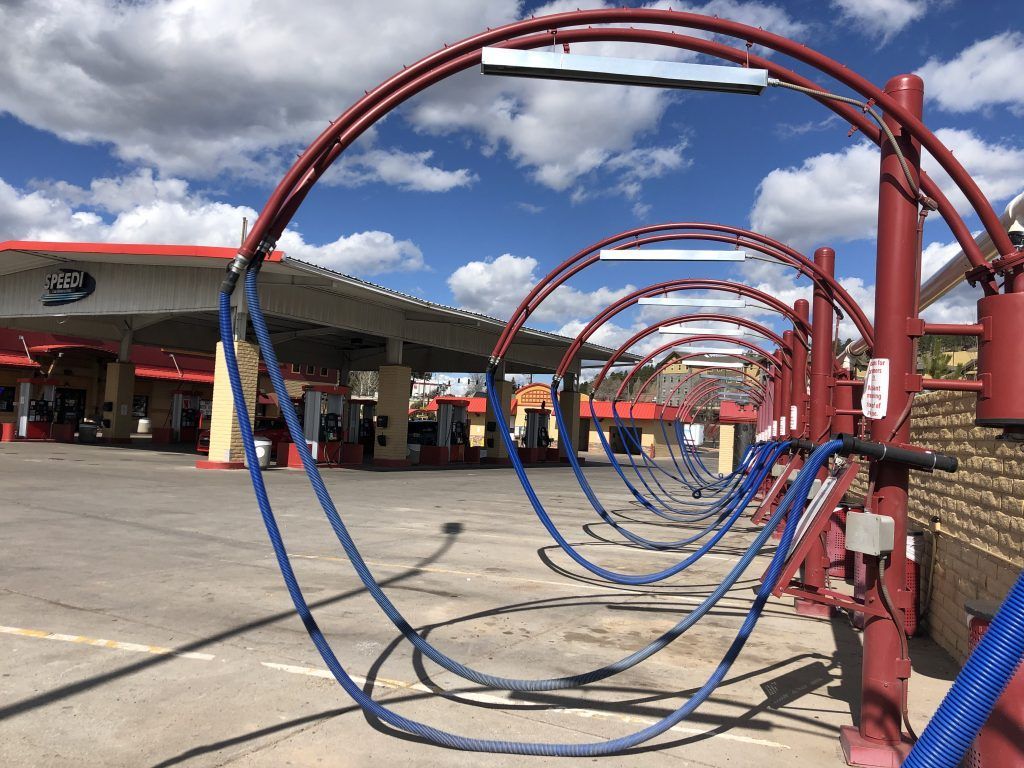 Car wash station with red arches and blue hoses, located at a gas station under a cloudy blue sky.