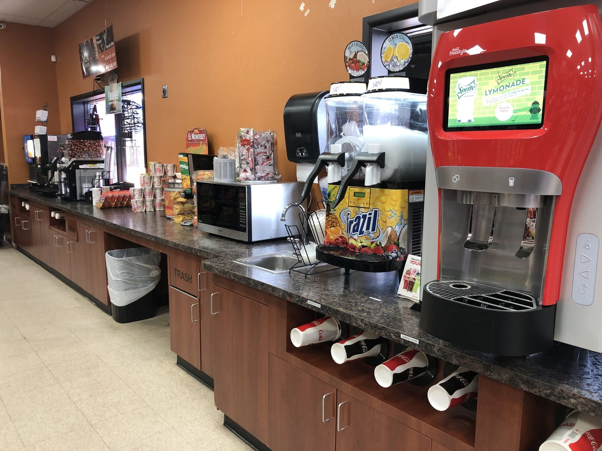 Interior of a convenience store with food and drink dispensers, microwave, snacks, and a trash can.