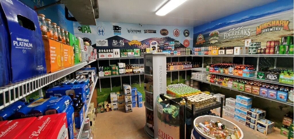 Inside a store, various beer brands and cases are stocked on shelves and coolers. A mural is on the wall.