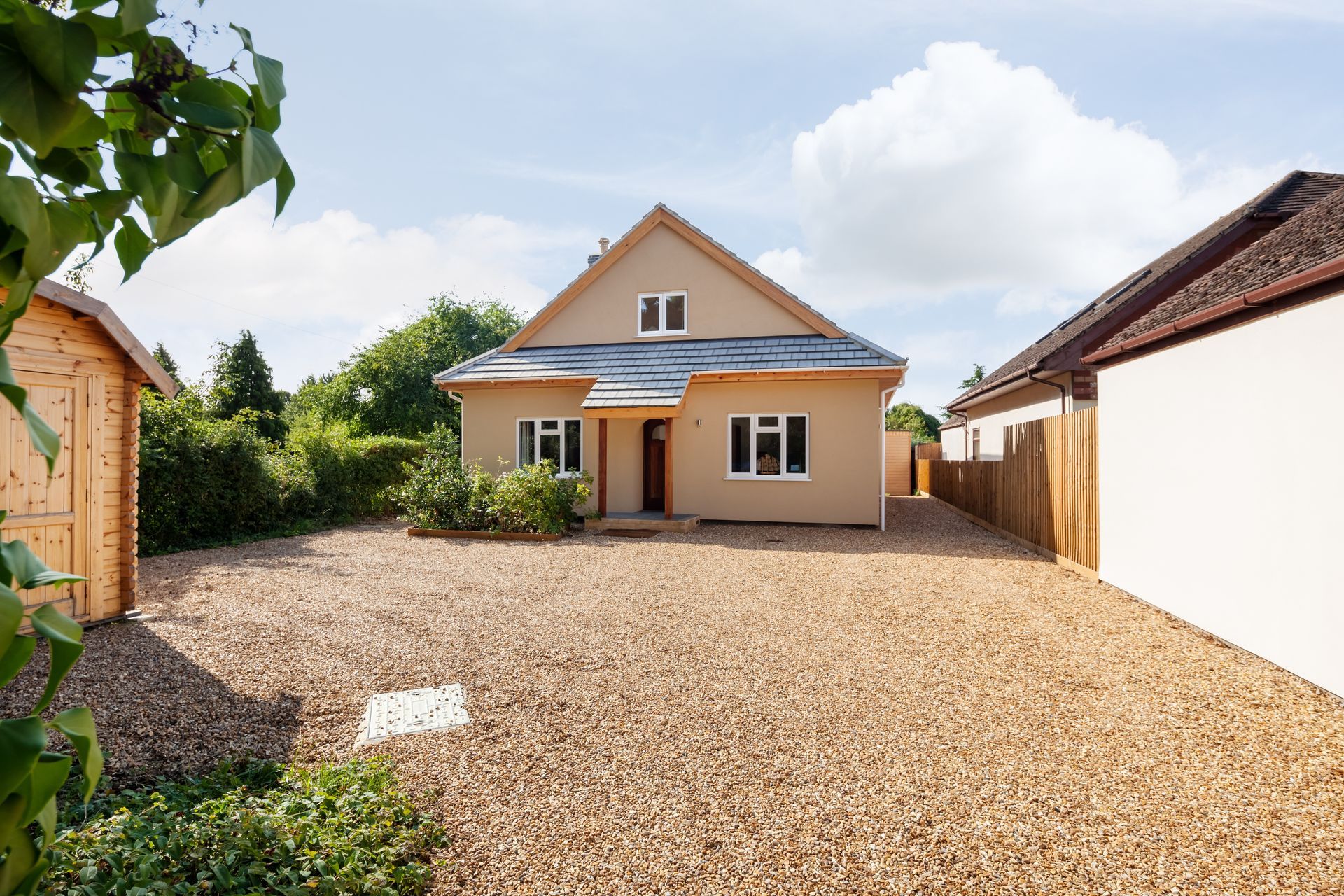 Single-story beige house with gravel driveway, white garage, and trees under a cloudy sky