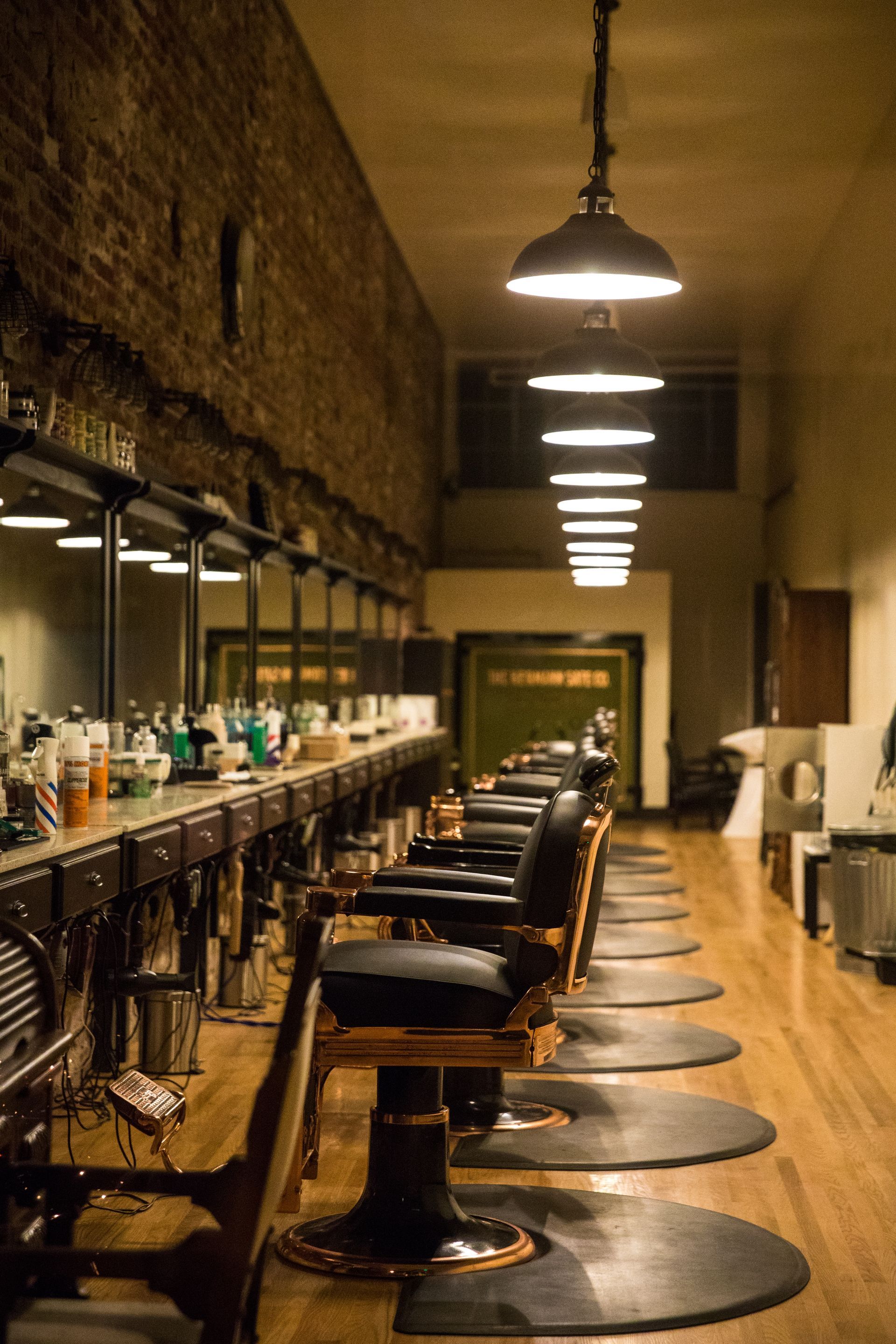 A row of vintage-style barber chairs in an old-fashioned shop with exposed brick walls, wooden floors, and hanging lamps.