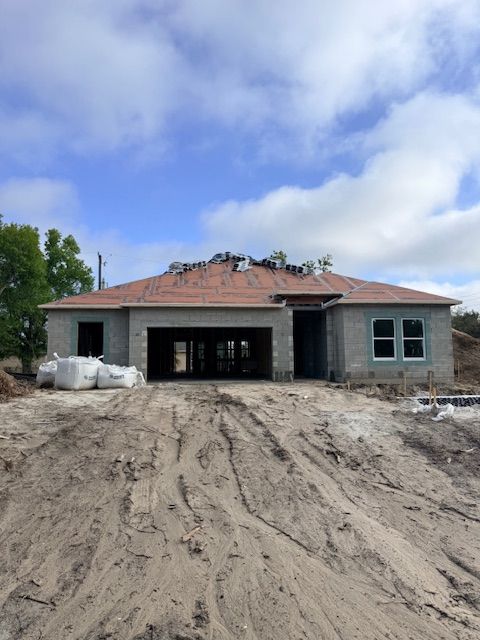 A newly constructed, single-story house with a partially shingled roof, unfinished concrete walls, and a dirt driveway.