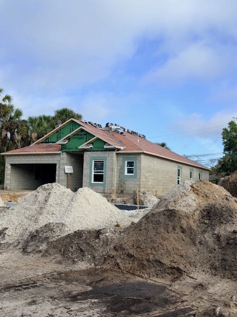 A house under construction with exposed gray concrete walls, green sheathing, and a shingled roof, surrounded by dirt piles.
