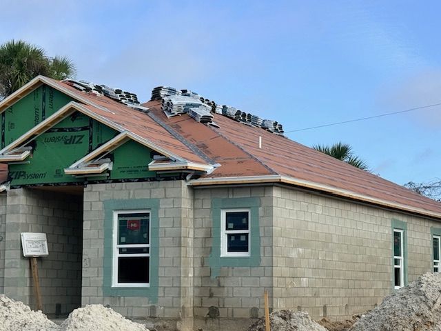 A partially constructed concrete block house with a red roof underlayment and green exterior sheathing under a blue sky.