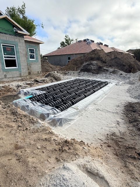 Construction site showing a rectangular underground plastic stormwater retention system in a sand pit next to a house.