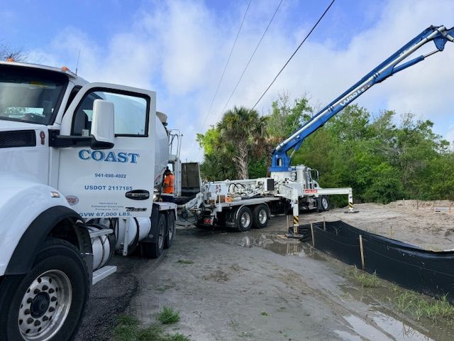 A Coast concrete pump truck with its boom arm extended over a construction site near trees and a black silt fence.
