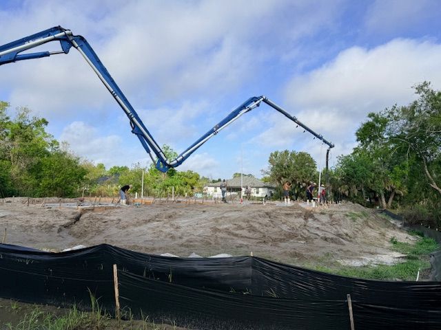 A blue concrete pump truck extends its boom arm over a dirt construction site with workers preparing the foundation.