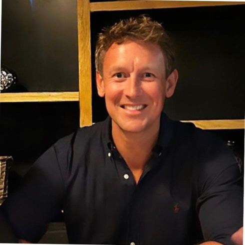 A person with short hair smiles at the camera, wearing a dark blue button-down shirt in a softly lit indoor setting.