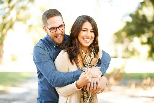 A smiling couple embraces outdoors in a park, with the man wrapping his arms around the woman from behind.
