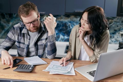 Two people sit at a table reviewing documents and using a laptop and calculator in a home kitchen setting.