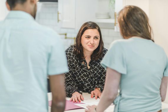 Three people in a brightly lit kitchen look at papers on a counter during a discussion.