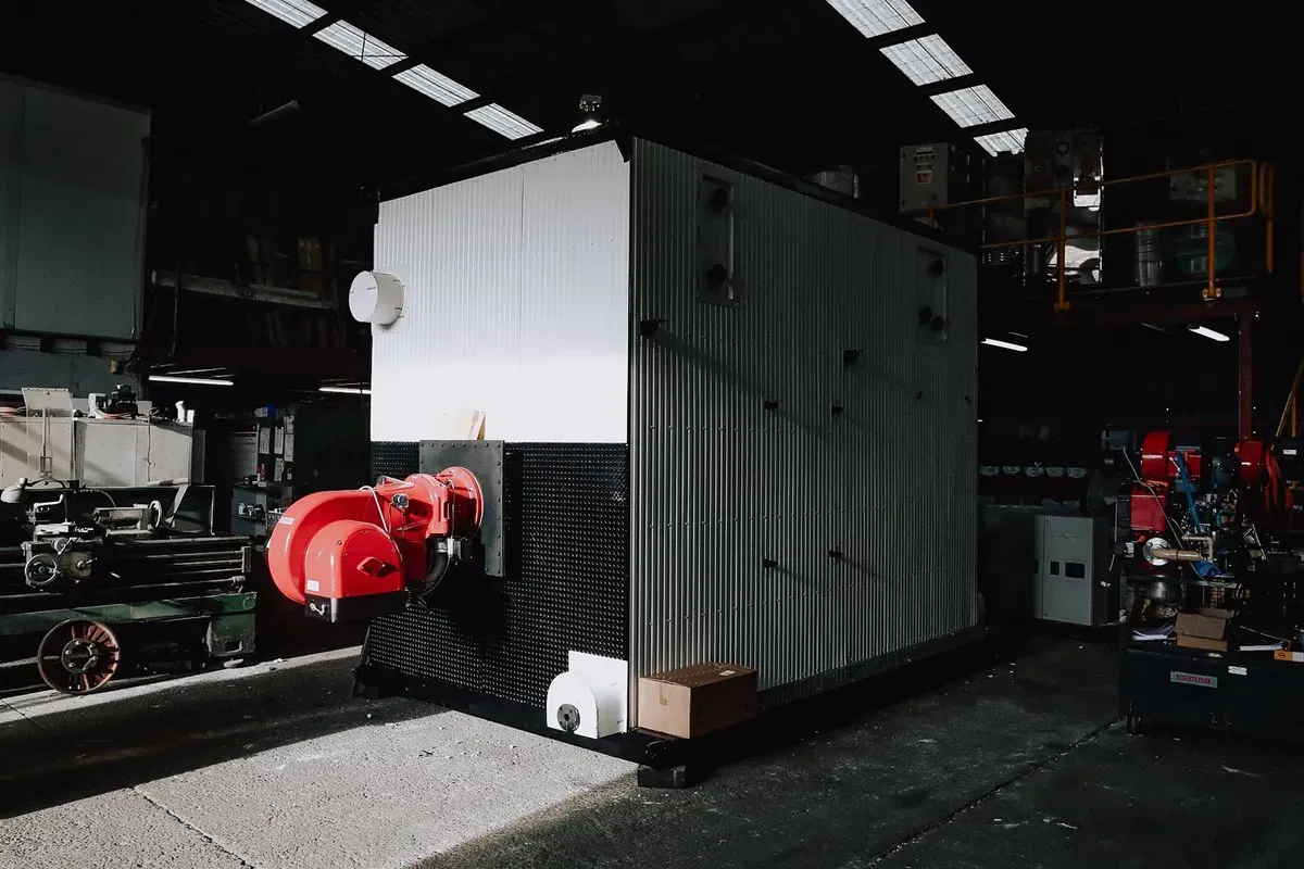 A large, white and grey industrial boiler with a red burner mechanism sits in a dimly lit, spacious warehouse facility.