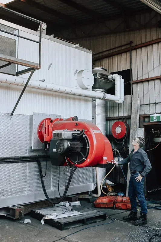 A person in work clothes inspects a large red industrial burner mounted on a white machine in an industrial workshop.