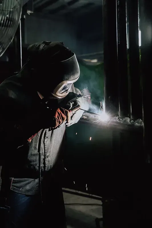 A person wearing protective gear and a welding helmet uses a torch to weld metal in a dimly lit workshop.
