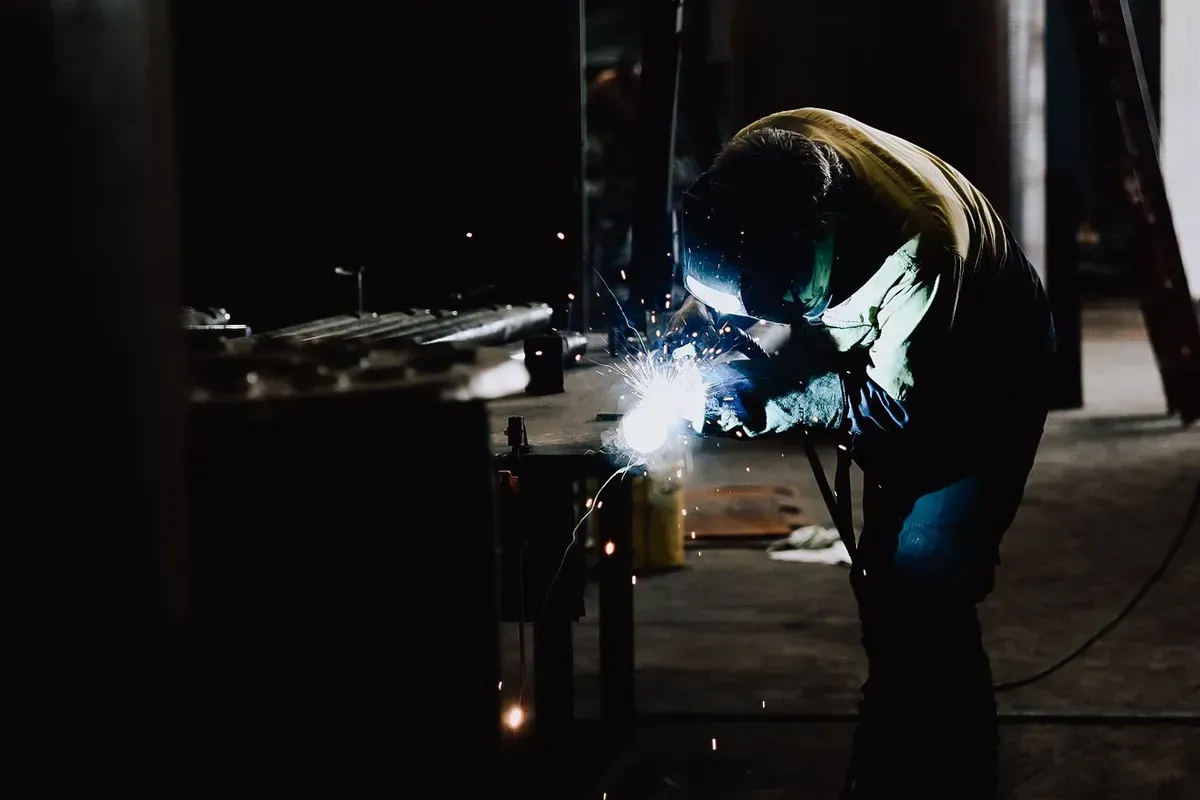 A welder wearing protective gear works on a piece of metal in a dim shop, creating bright sparks during the process.
