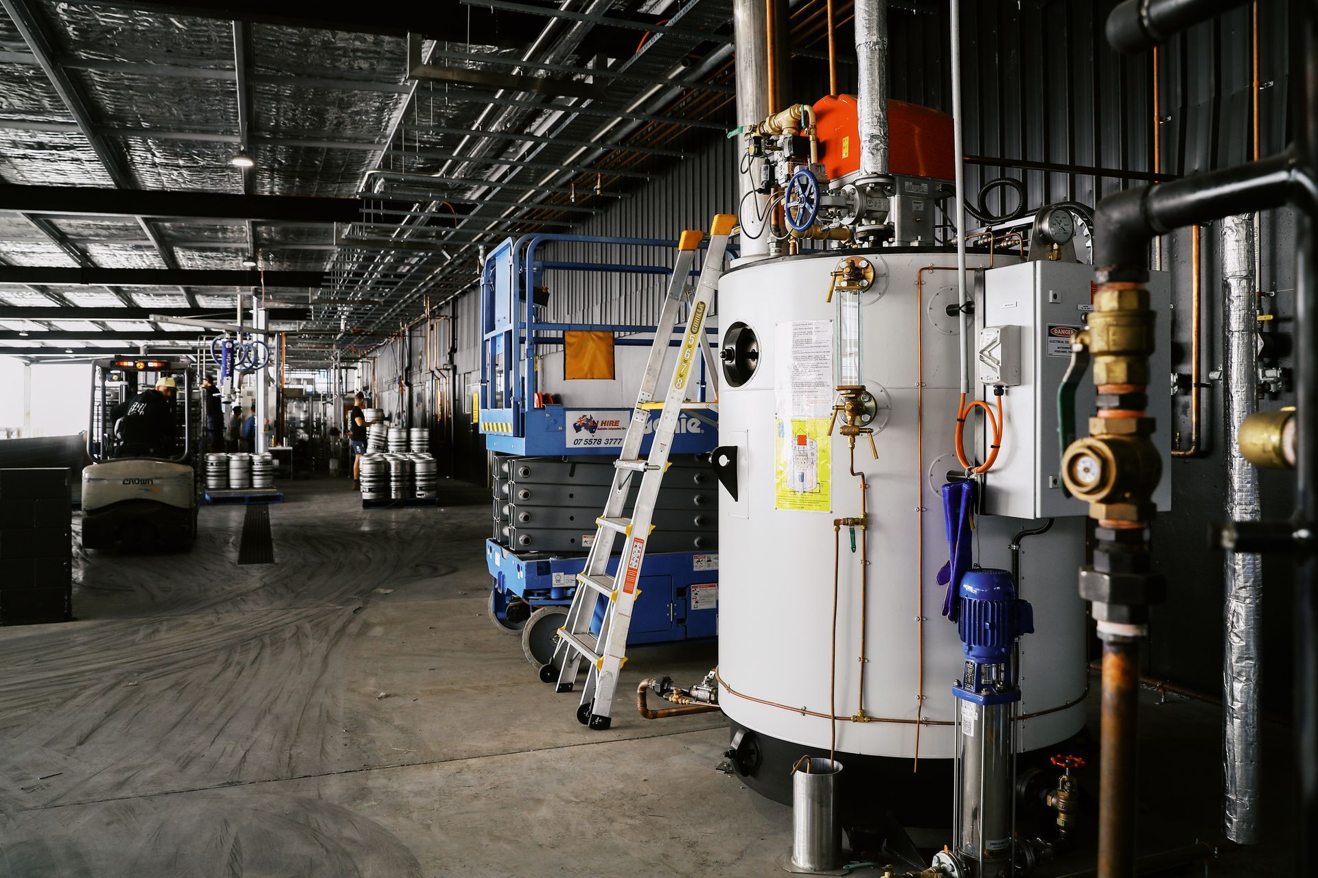 Industrial facility with a large white boiler, a blue scissor lift, metal kegs, and a forklift in the background.