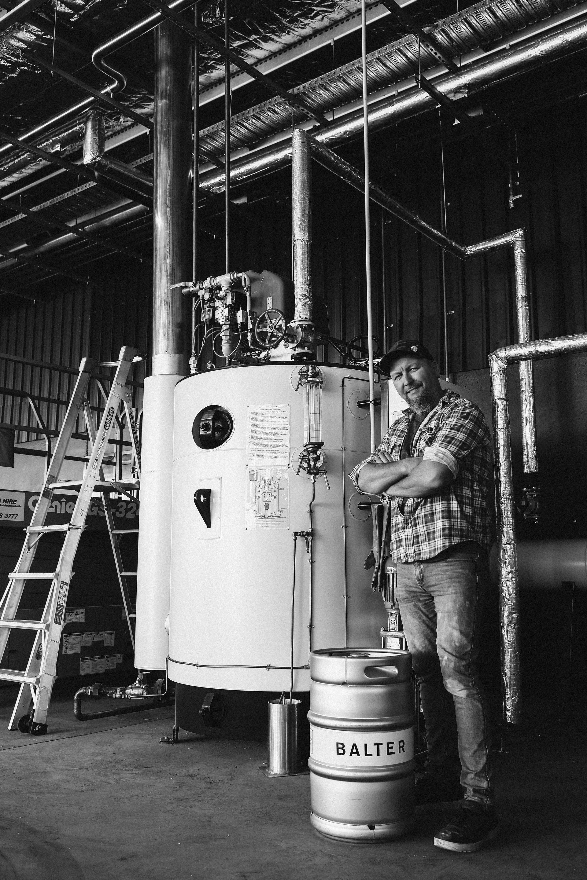A person stands with arms crossed next to a large industrial boiler and a keg in a warehouse setting.
