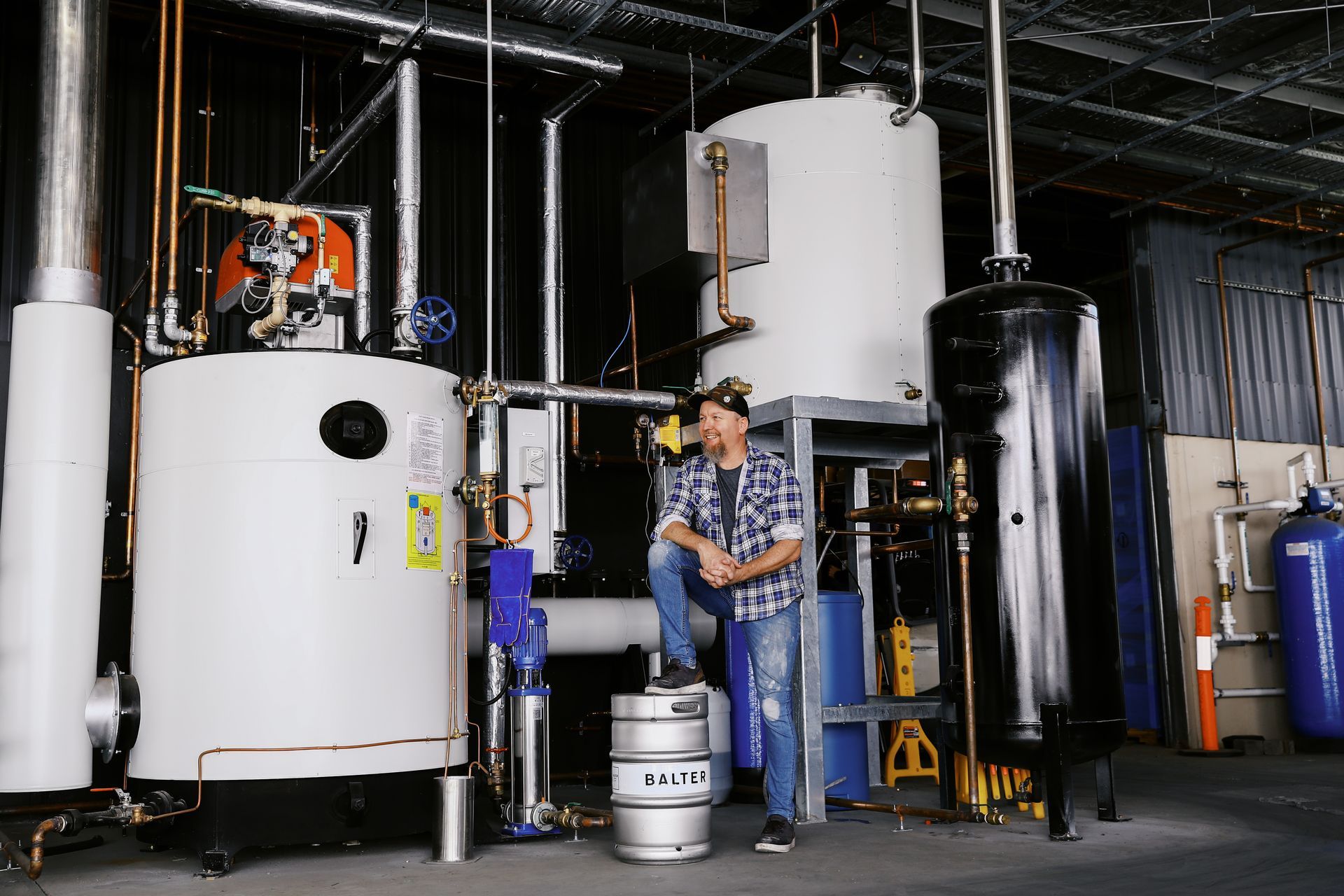 A person wearing a plaid shirt stands in an industrial facility next to large, white and black metal tanks and a keg.