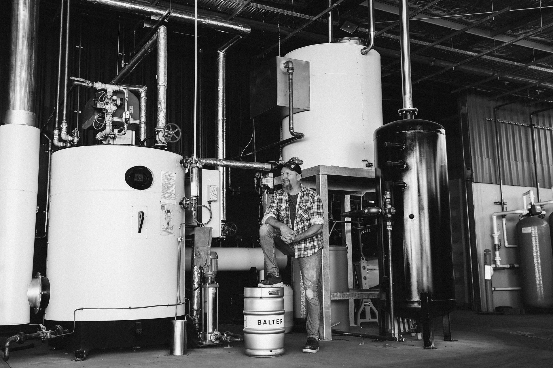 A worker standing beside a stainless steel keg in an industrial room with large boilers and tanks.