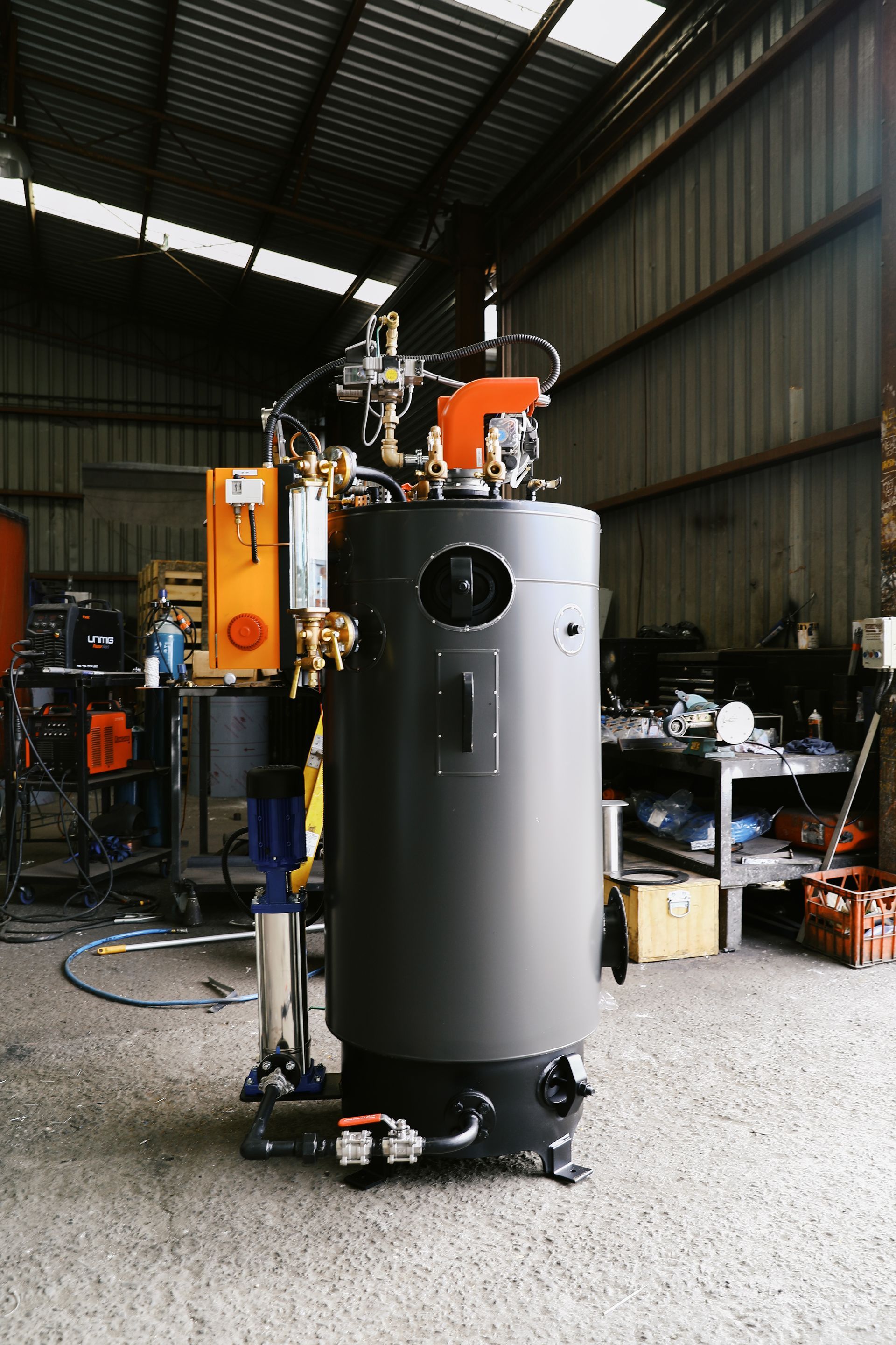 A vertical industrial boiler with a grey cylindrical body, orange components, and attached pipes in a factory workshop.
