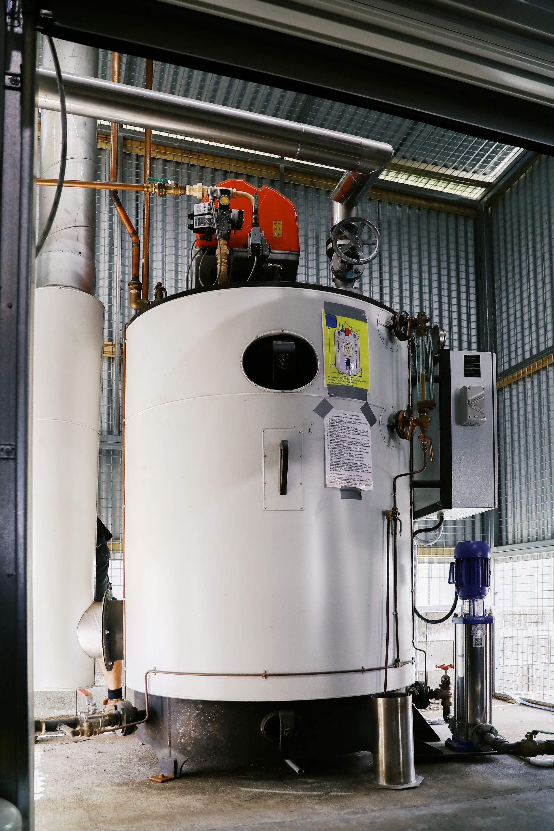 A white industrial boiler stands in a room with corrugated metal walls, featuring a top-mounted orange burner and pump.